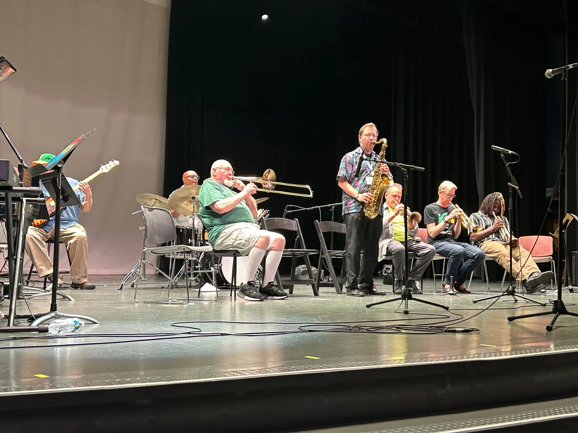 A standing saxophonist plays onstage alongside seated trombone, trumpet, guitar, and rhythm players.