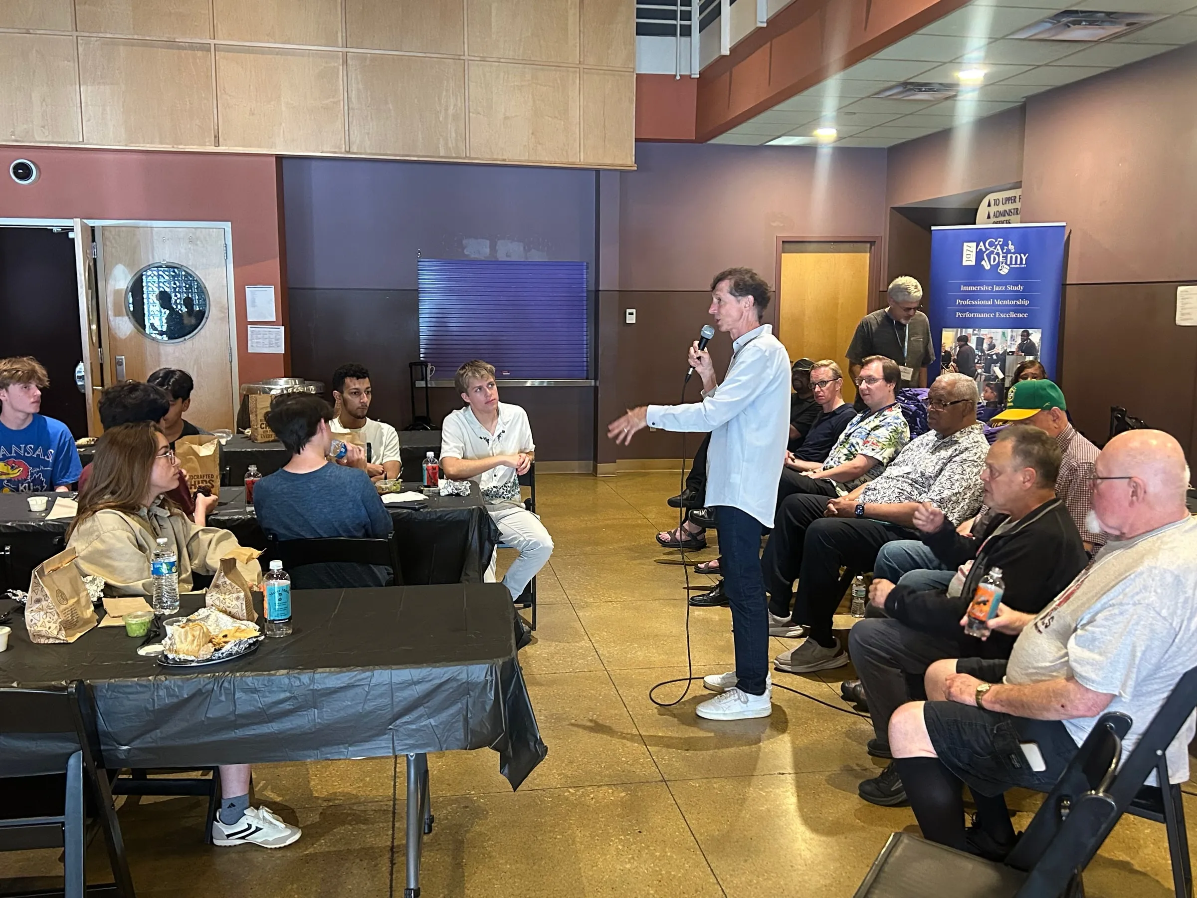 Campers and adults seated around tables listen to a speaker with a microphone during a luncheon learn.