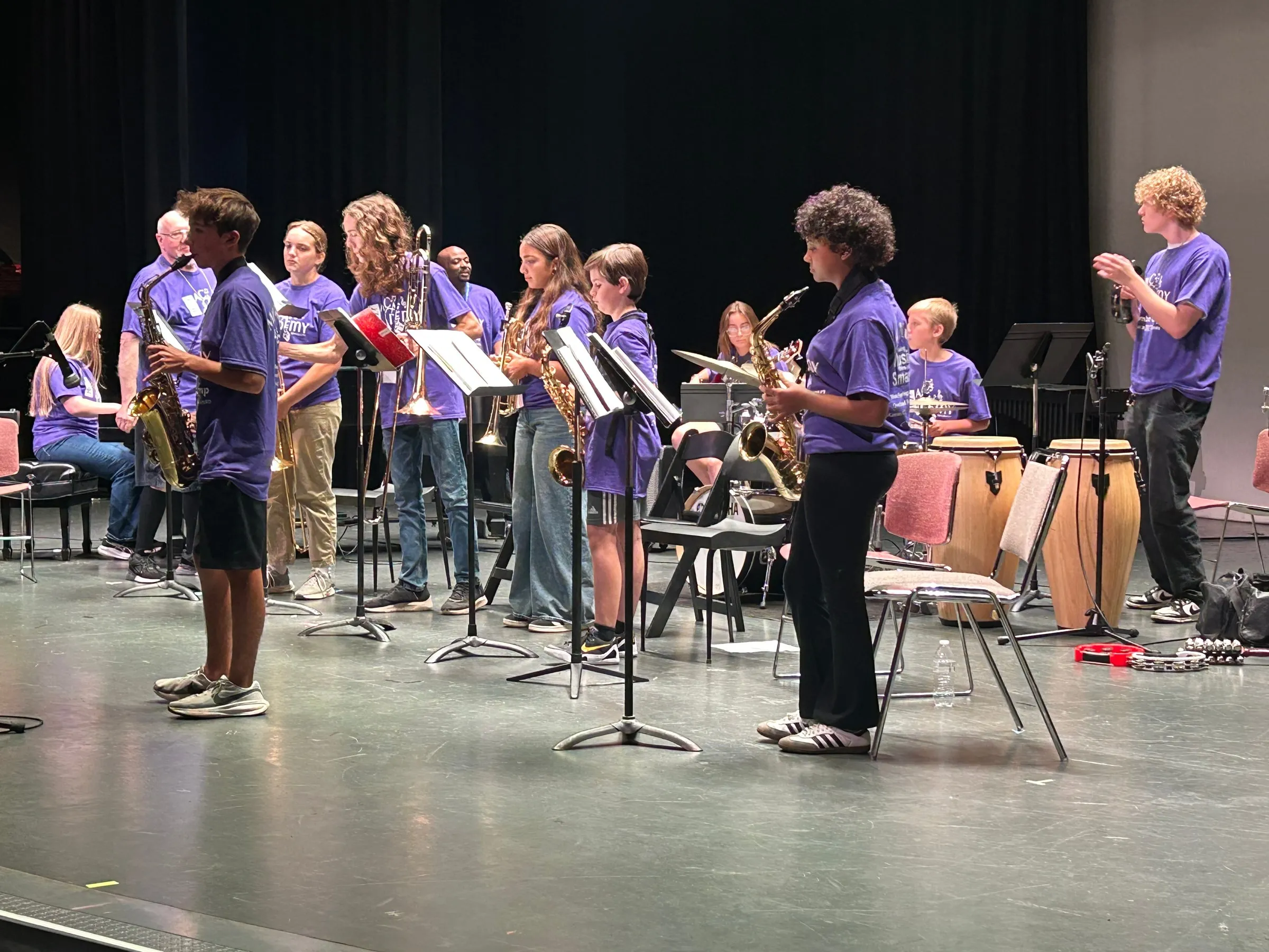 Young musicians in purple camp shirts applaud onstage with their instruments and music stands.