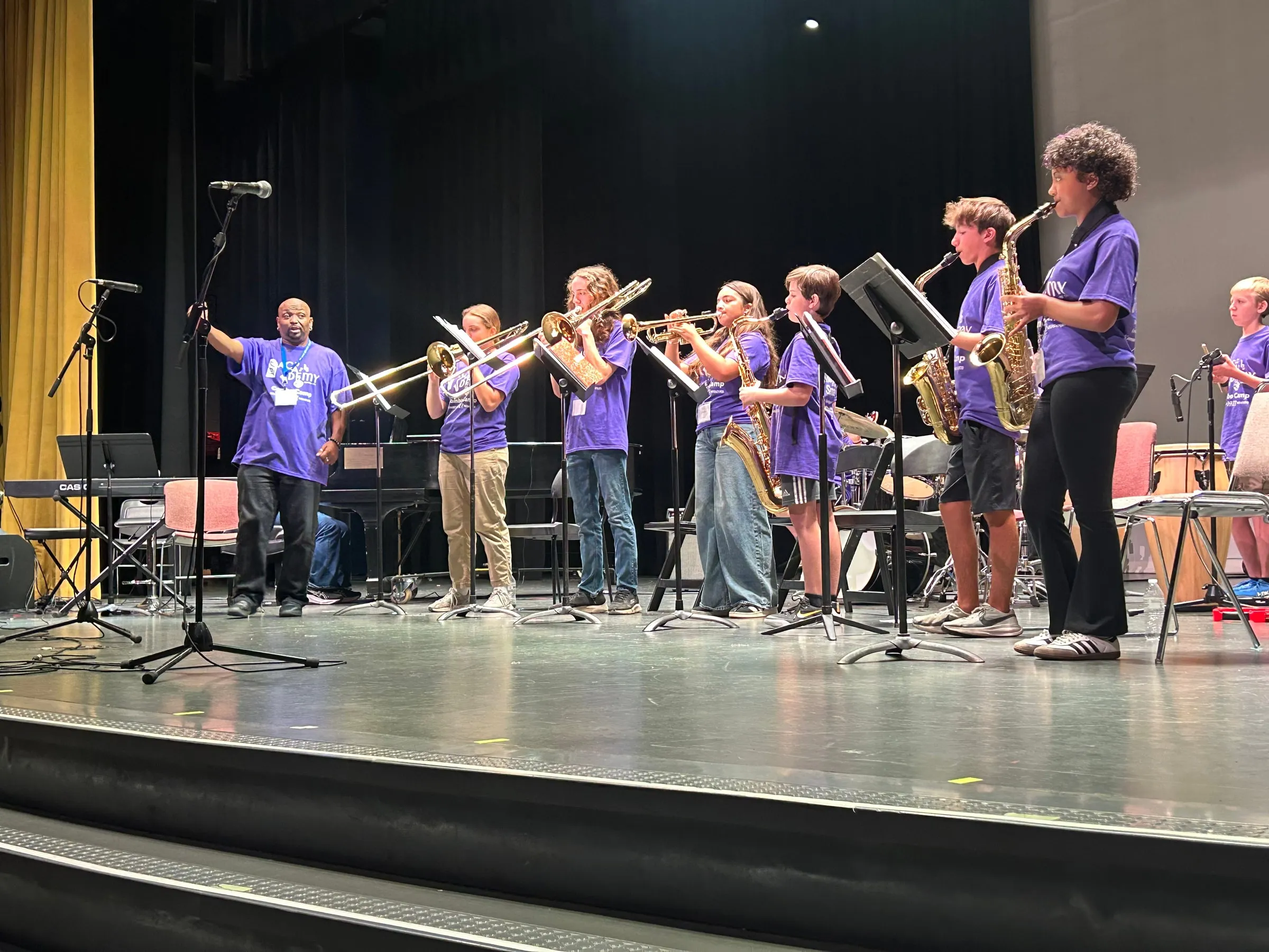 A director cues a standing ensemble of young brass and saxophone players in purple camp shirts onstage.
