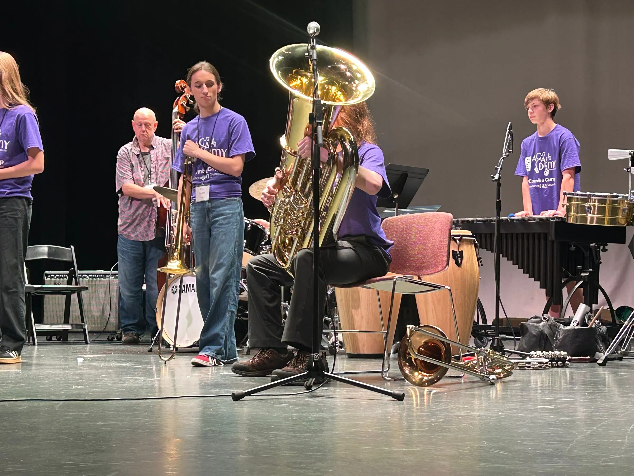 A seated tuba player performs beside a saxophonist, with a percussionist at the mallet instrument behind them.
