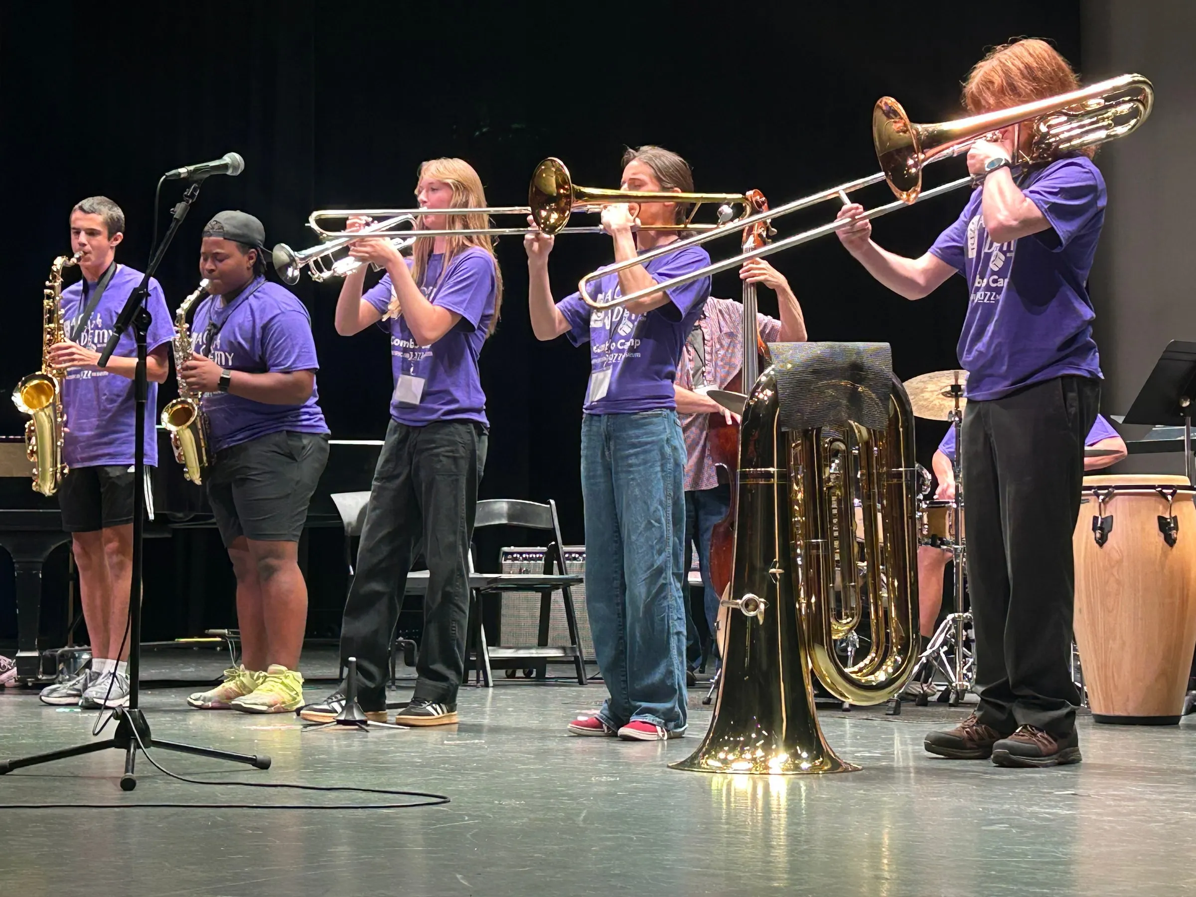 A row of saxophone, trumpet, and trombone players performs together in purple camp shirts.
