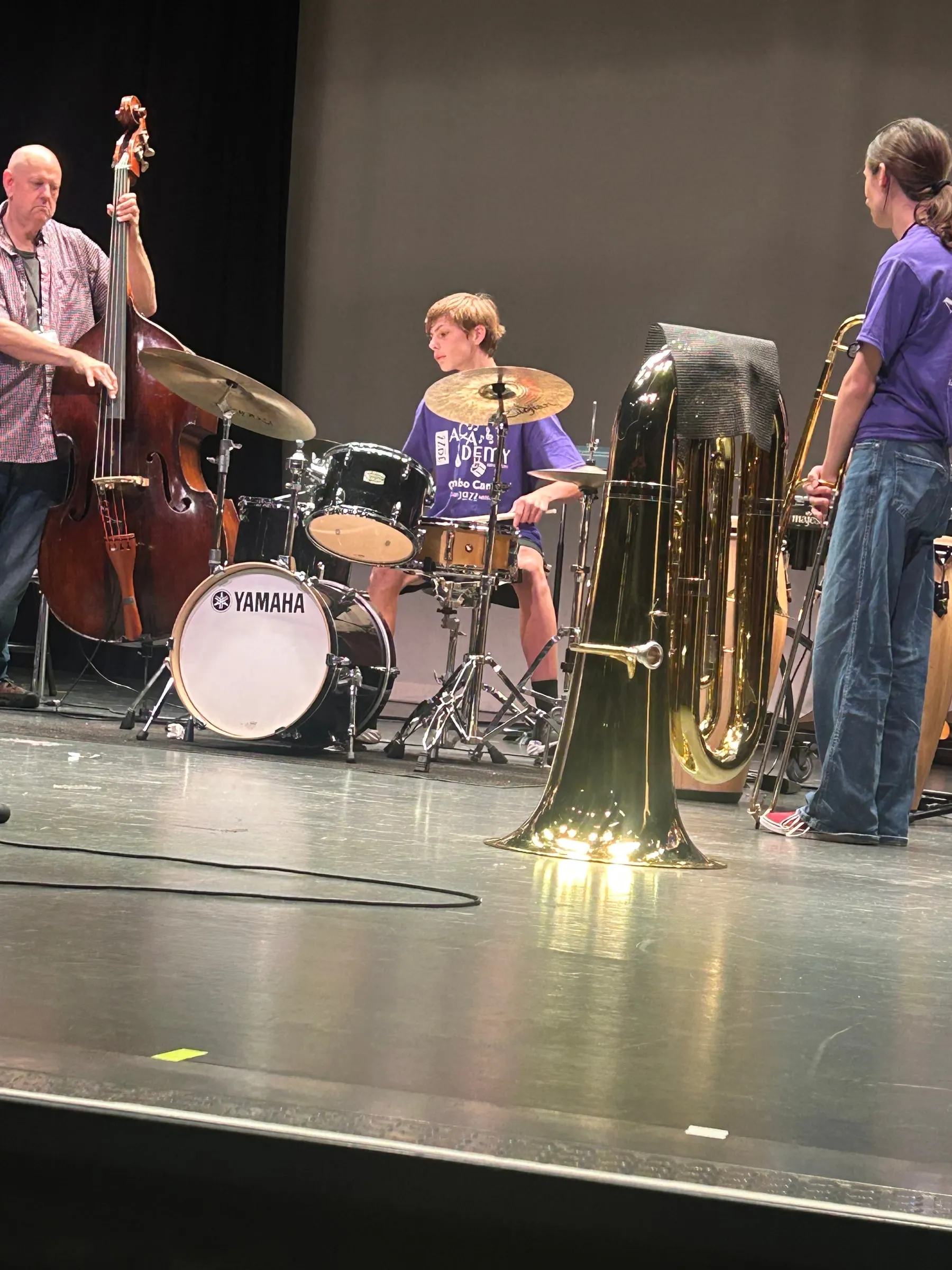 A student plays the drum kit beside an upright bassist, with a tuba in the foreground.