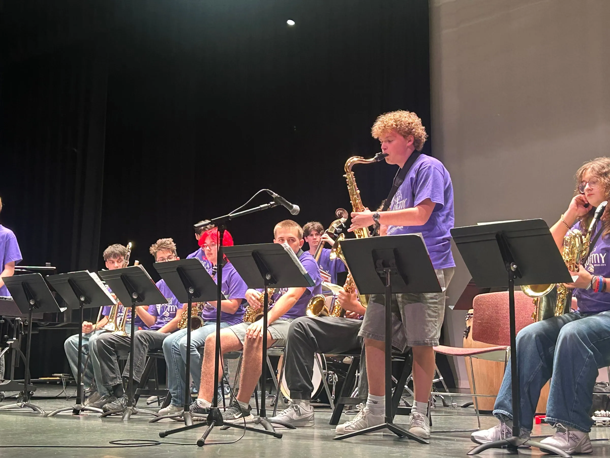 A standing saxophone soloist plays into a microphone in front of the seated ensemble.