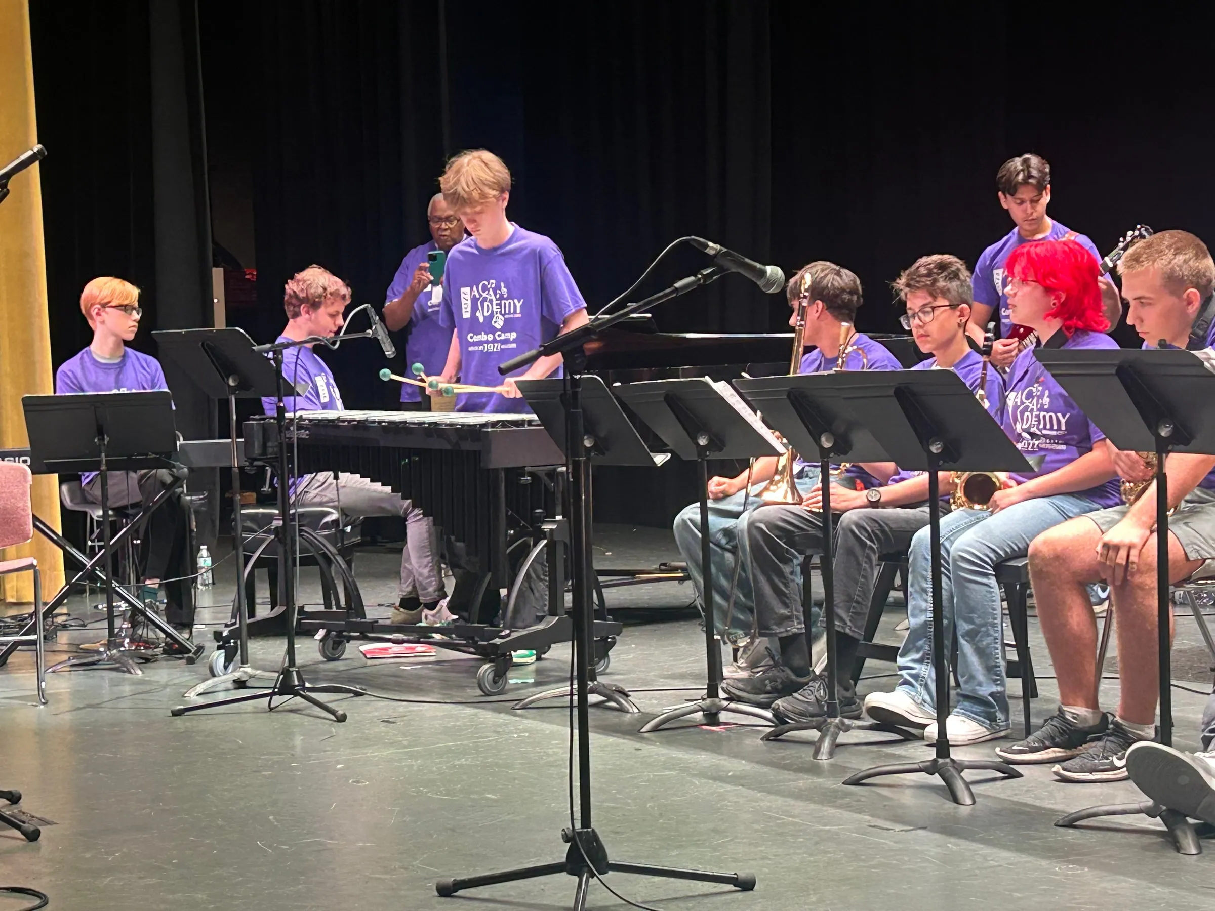 A student plays mallet percussion behind a microphone, surrounded by seated ensemble members.