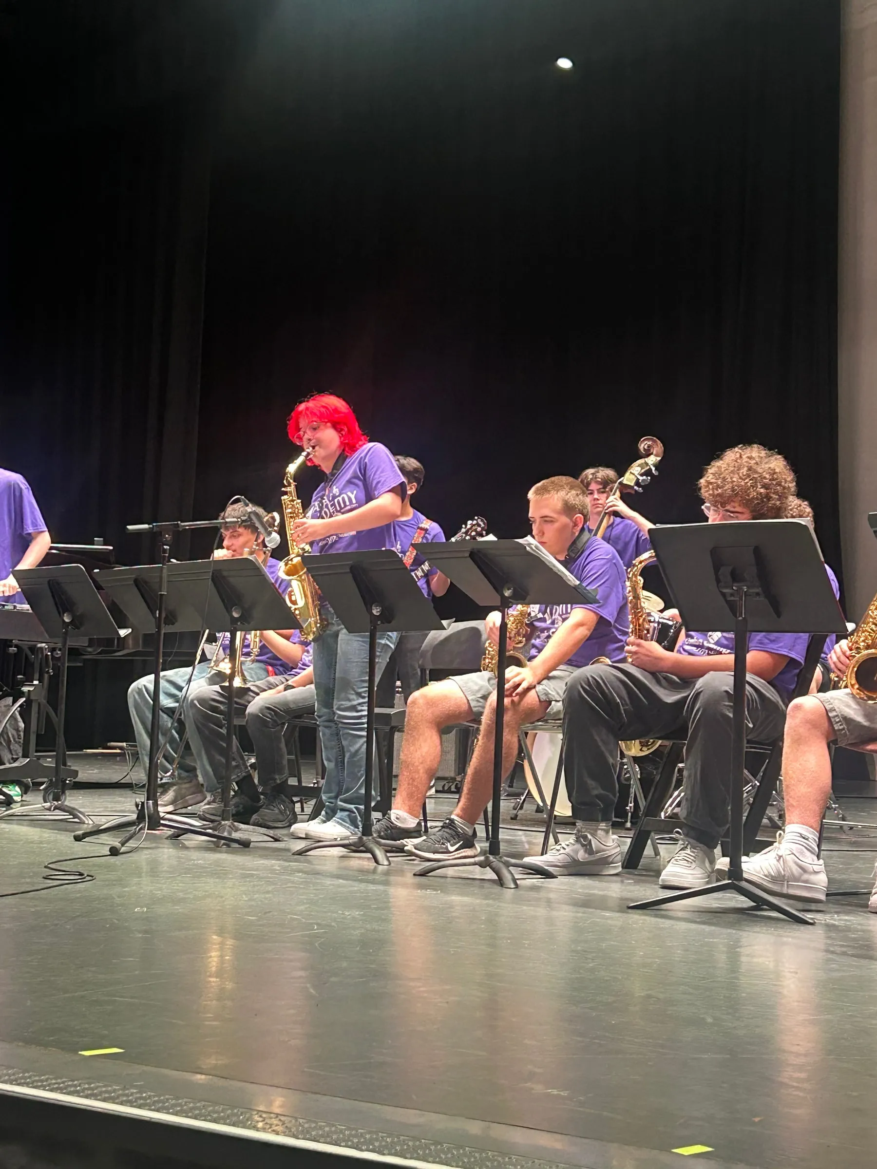 A saxophone player stands to perform a solo among rows of seated musicians and music stands.