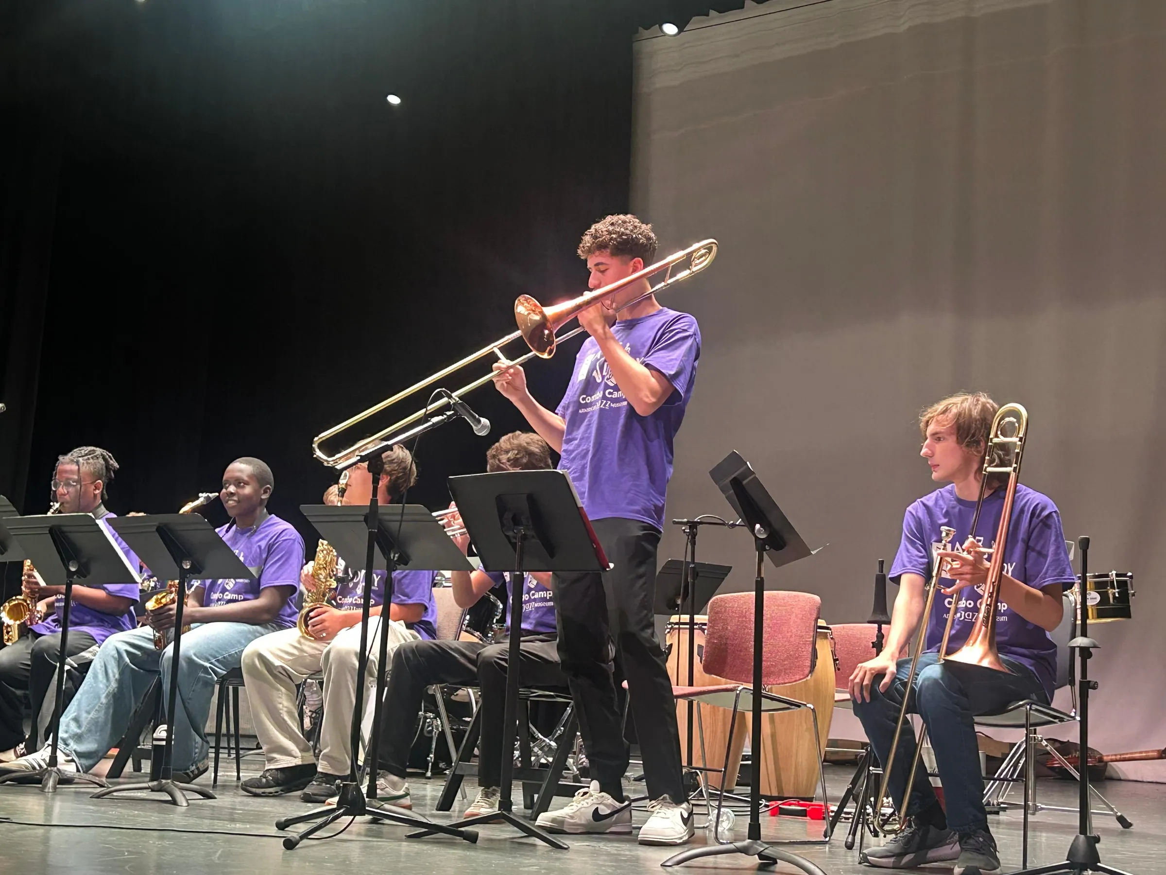 A trombone soloist plays with the slide extended toward a microphone beside the seated horn section.