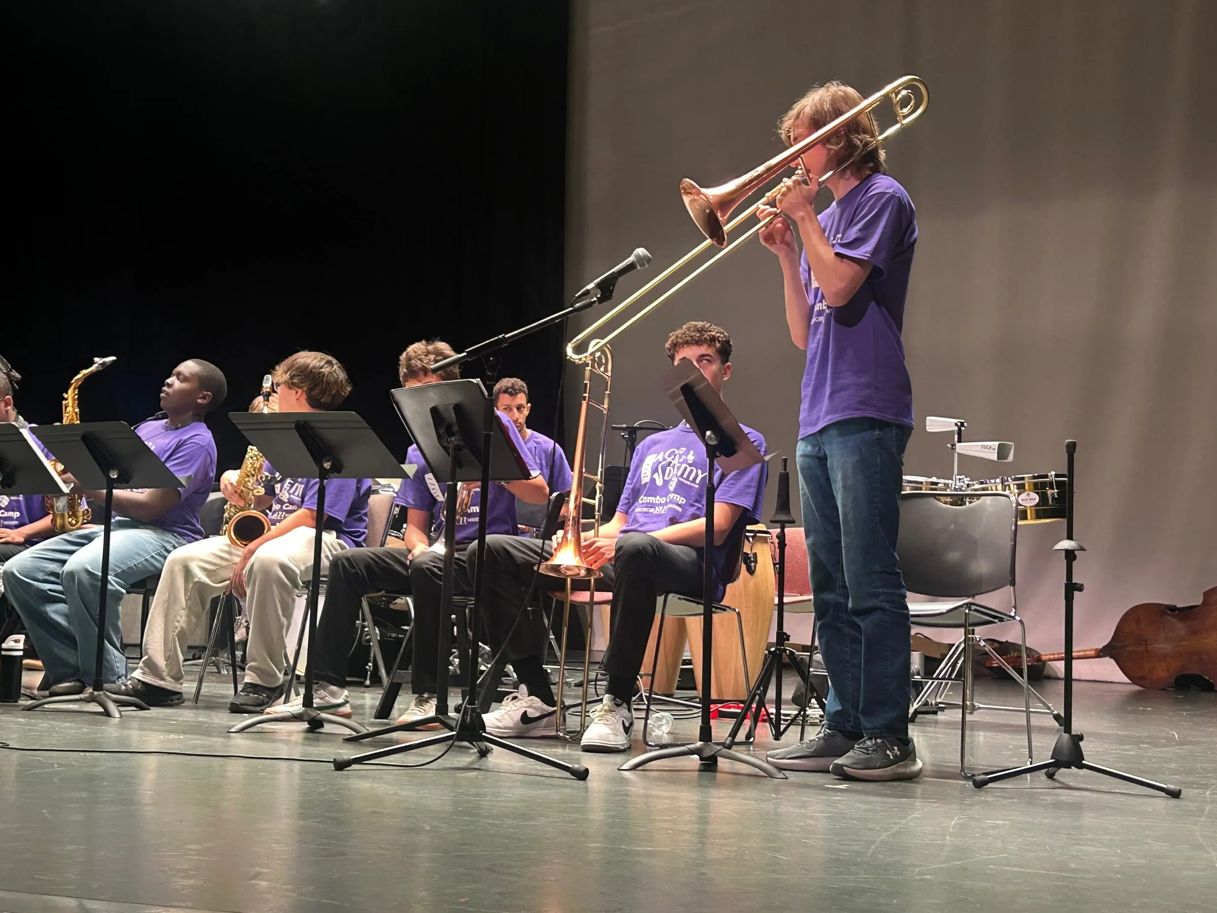 A standing trombone player performs at the front of the stage while the seated ensemble looks on.