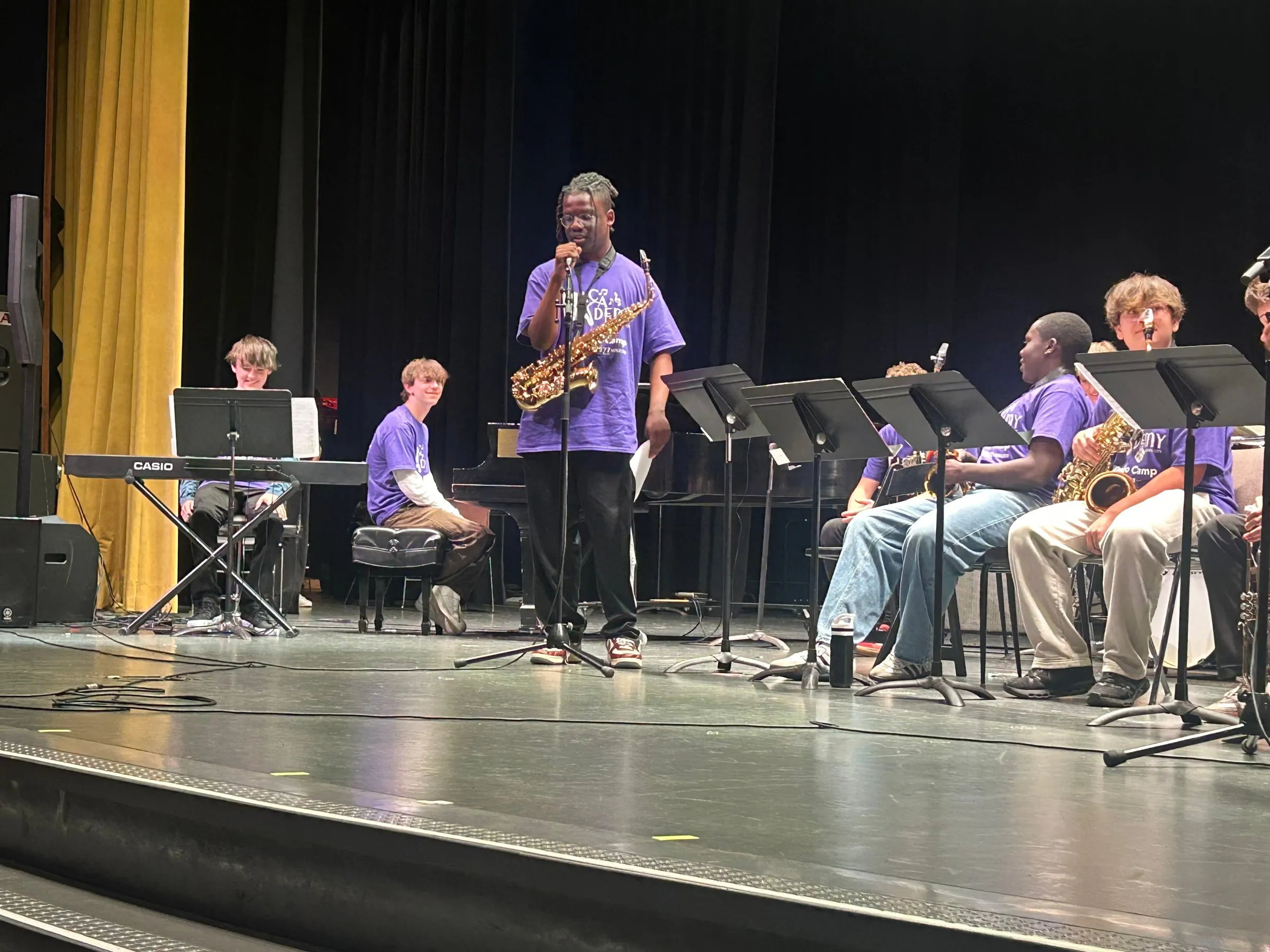 A student holding a saxophone speaks into a microphone beside the seated ensemble and pianist.