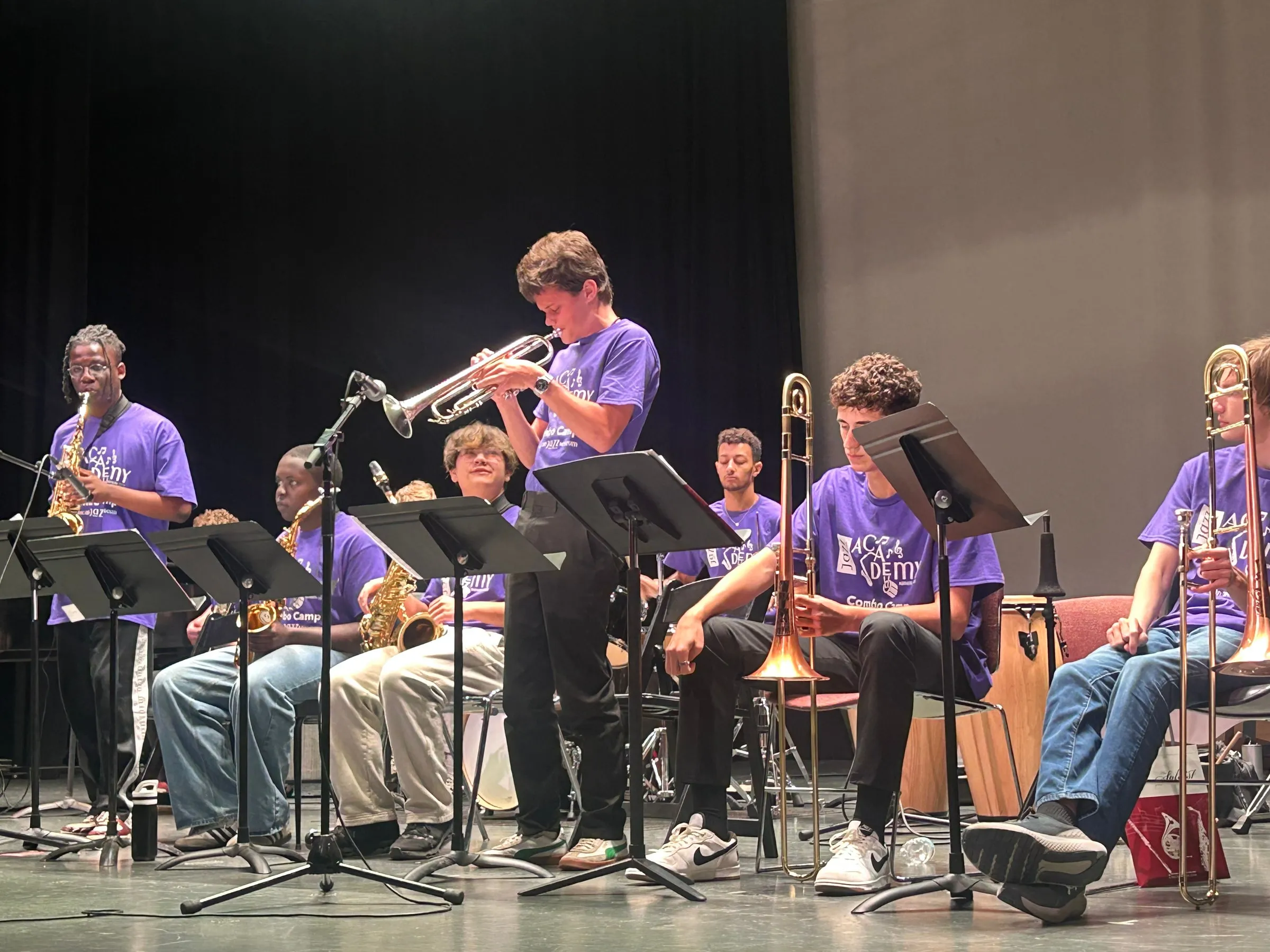 A trumpet soloist plays into a microphone among seated saxophone and trombone players.
