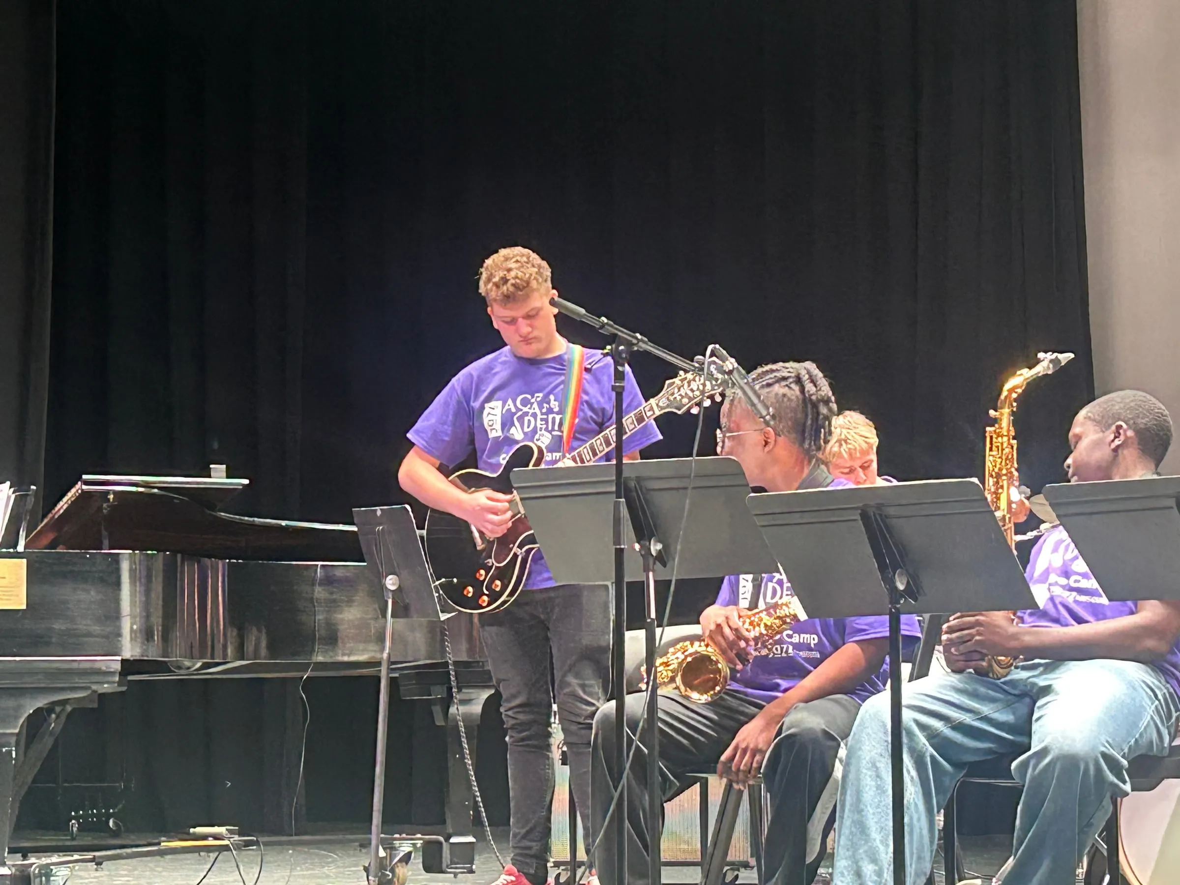 A student plays electric guitar behind the seated saxophone section near the grand piano.