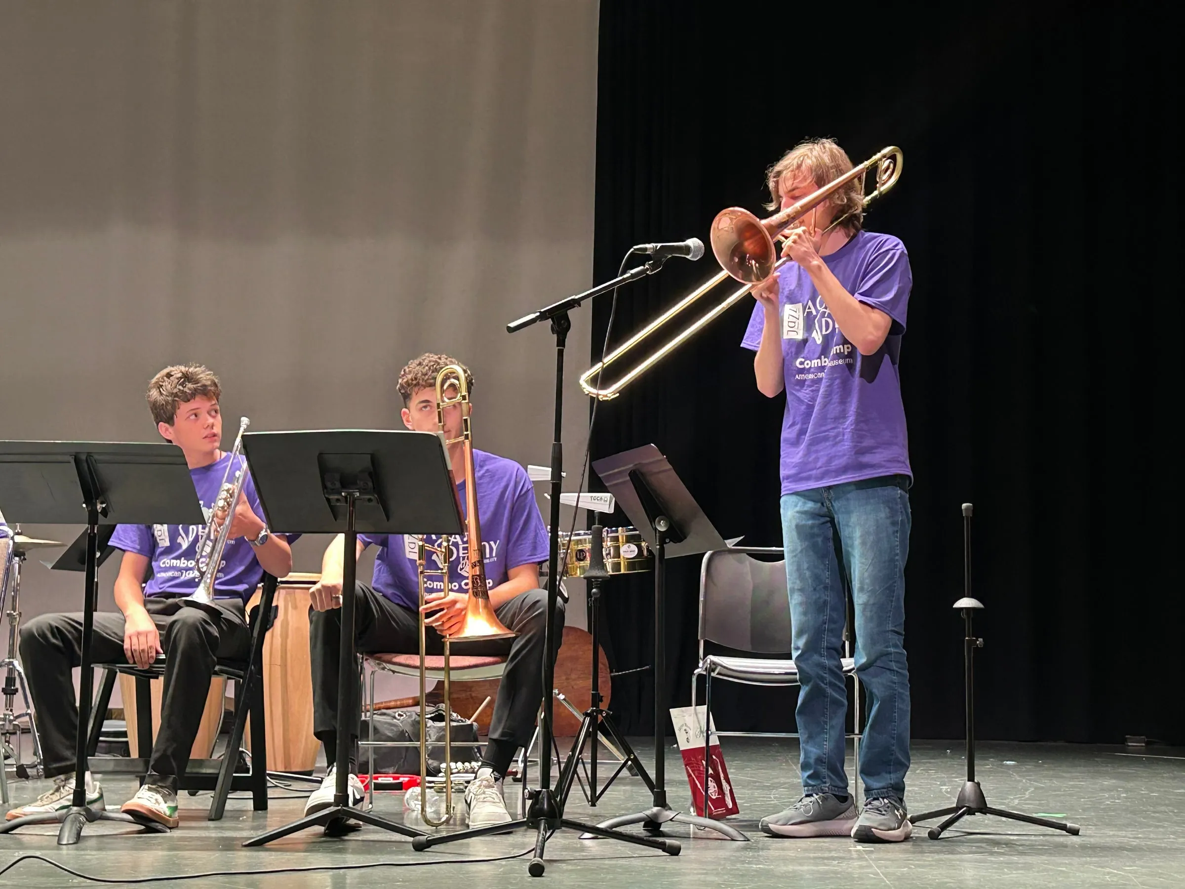 A trombone soloist stands at a microphone while two seated trombonists watch from behind music stands.