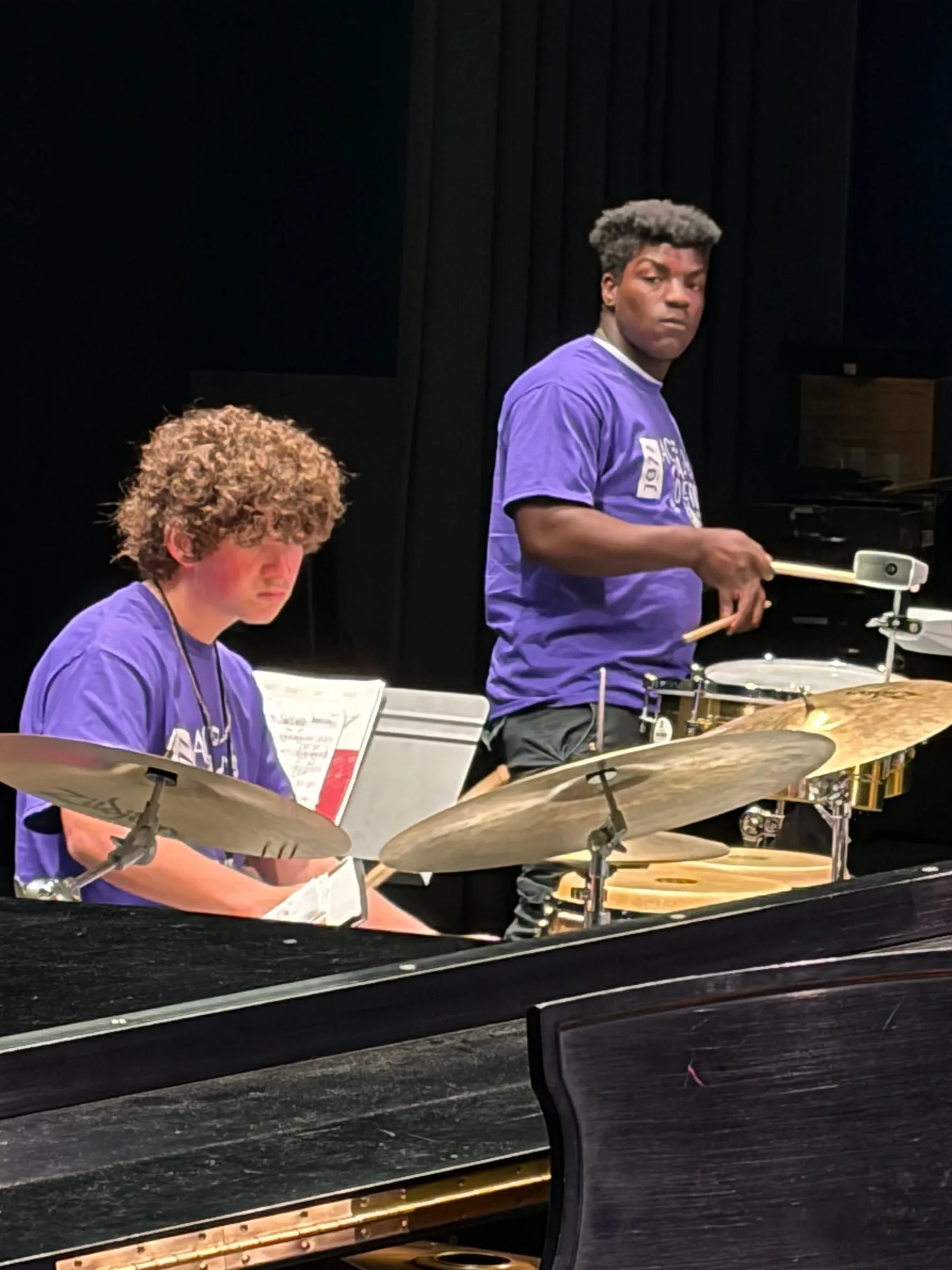A drummer and a standing percussionist perform together behind cymbals, reflected in a glossy piano lid.