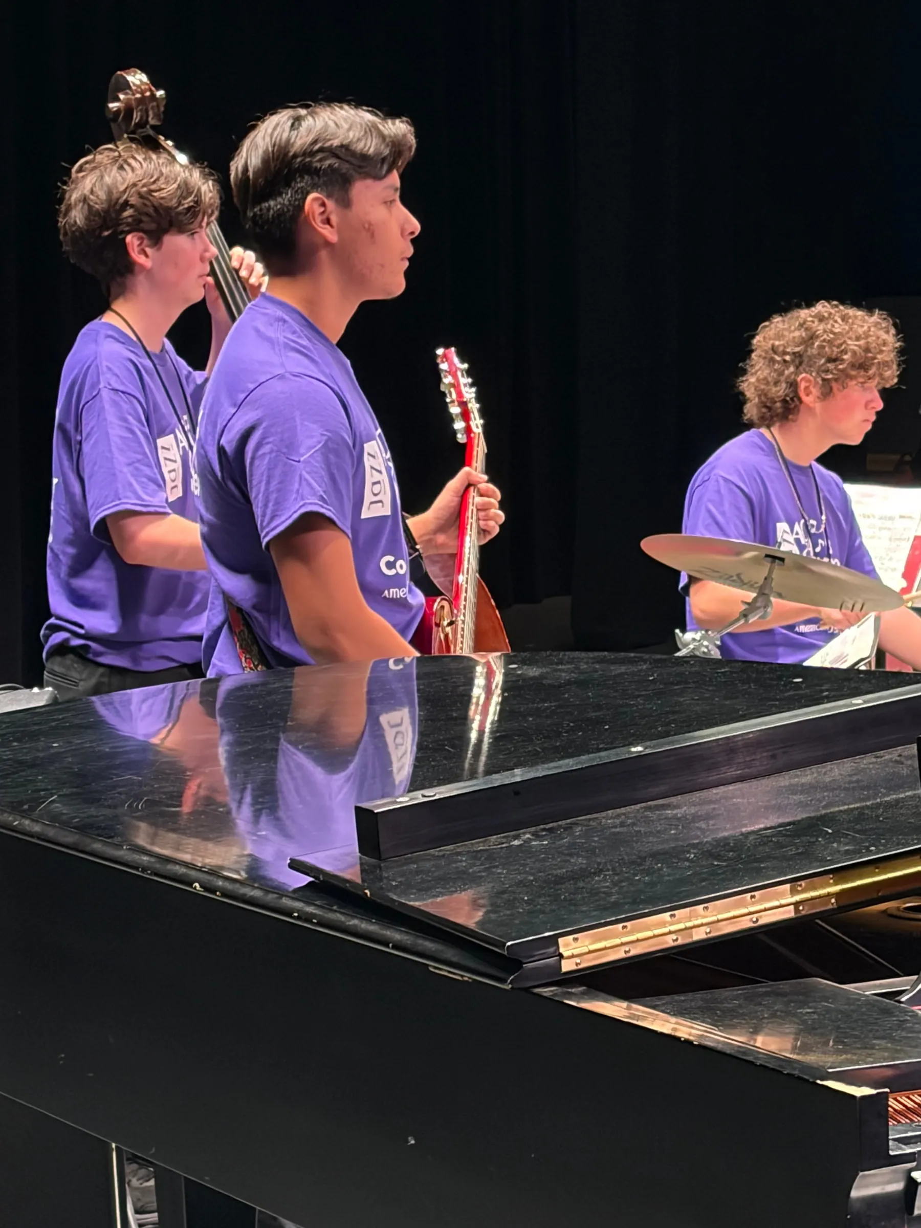 A side-stage view of guitar, upright bass, and drums with the musicians reflected in the piano lid.